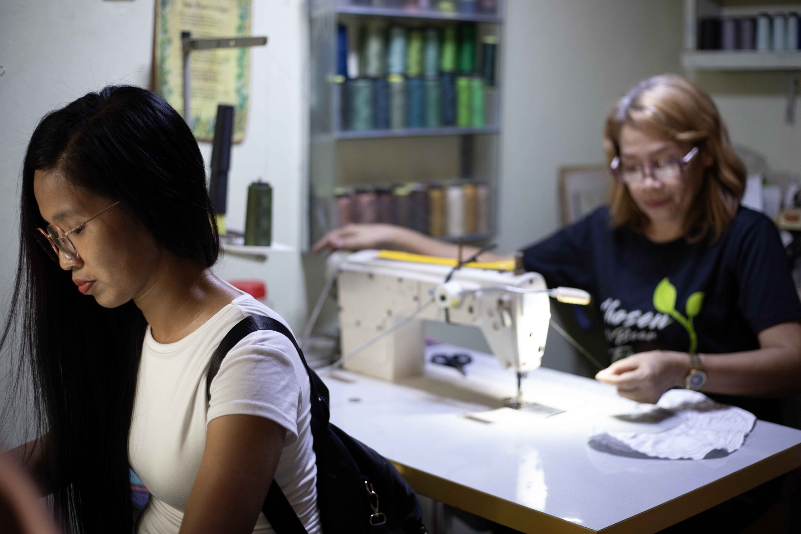 Two women work in a sewing room, one focused on a sewing machine while the other prepares fabric.