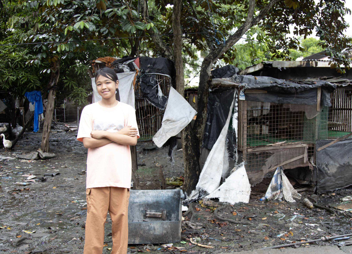 A young girl stands confidently in a rundown area with makeshift shelters and debris in the background.