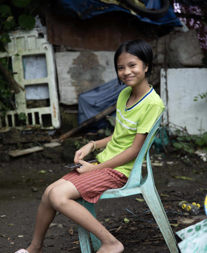 A young girl in a green shirt sits on a plastic chair, smiling in a cluttered outdoor environment.