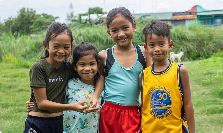 Four smiling children standing together outdoors in a grassy area