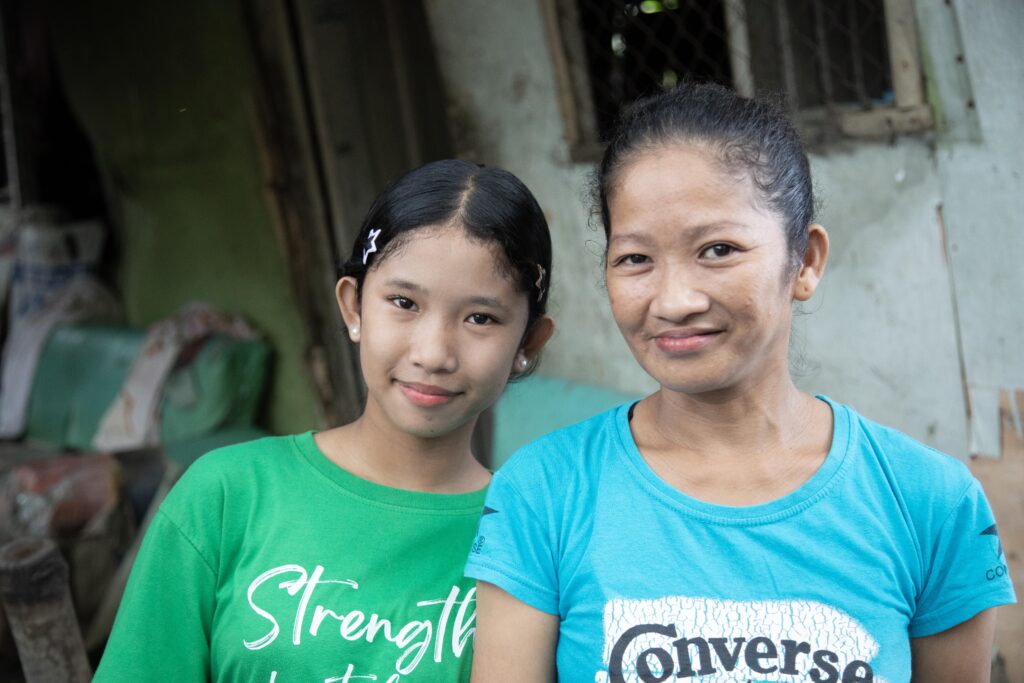 Two smiling women, a young girl in a green shirt and an adult in a blue shirt, pose together in a rustic outdoor setting.