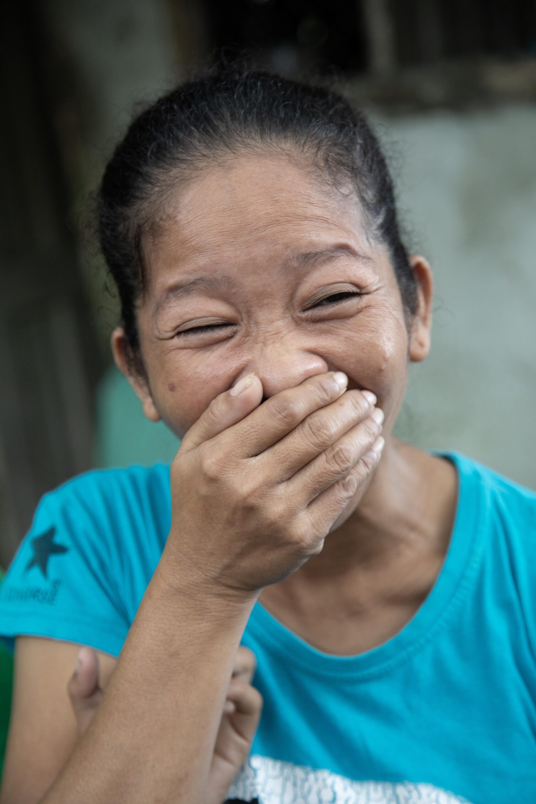 A woman in a blue shirt laughs joyfully, covering her mouth with her hand, conveying happiness and warmth.