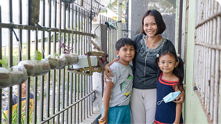 A woman stands with a boy and girl on a balcony, smiling, with birds in cages visible in the background.