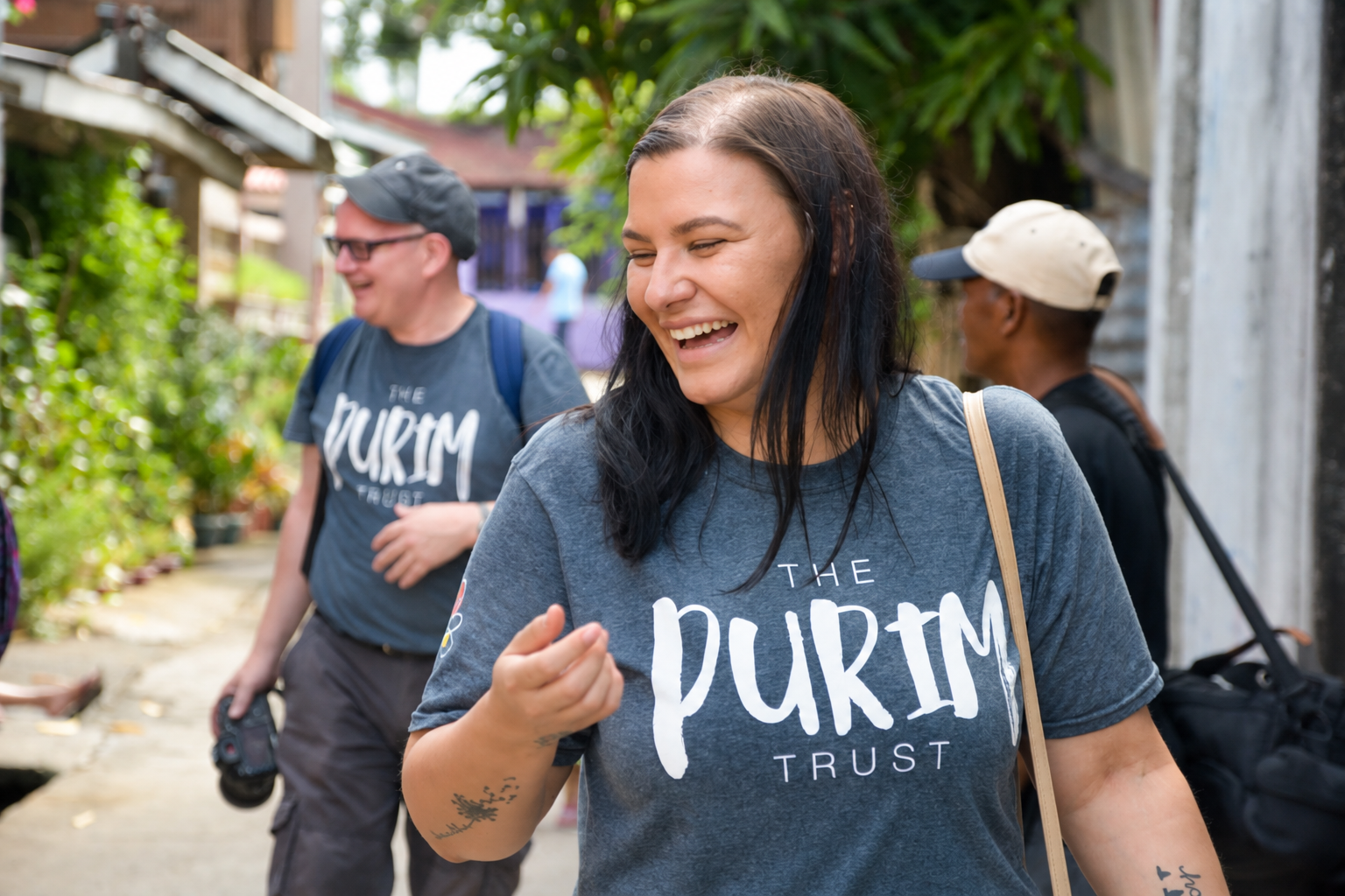 Woman laughing outdoors with two people behind her; they wear T-shirts reading “The Purim Trust”